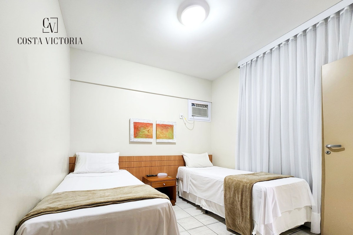 Habitación de hotel con dos camas individuales, cabecero de madera, mesita de noche pequeña y ventana con cortinas blancas.