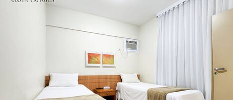 Habitación de hotel con dos camas individuales, cabecero de madera, mesita de noche pequeña y ventana con cortinas blancas.