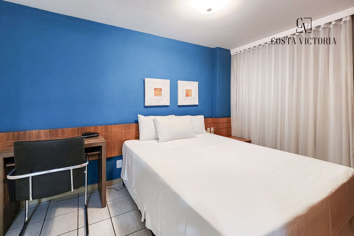 A hotel room with a large bed, a desk, and two framed pictures on the wall.
