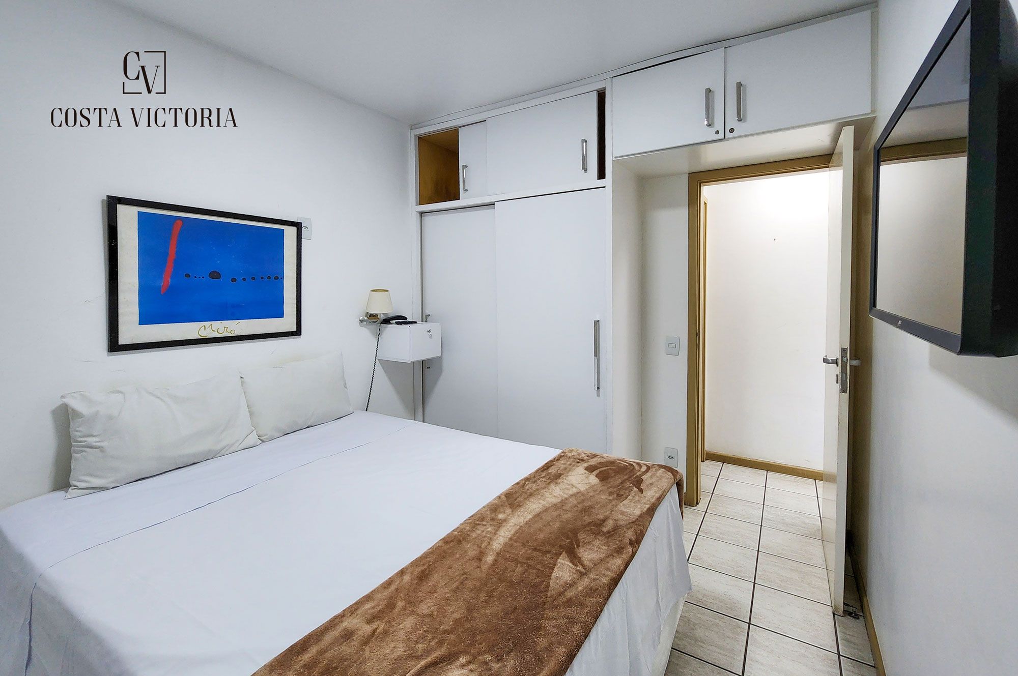 Habitación de hotel con cama, escritorio, silla, televisor y puerta.