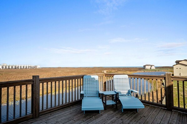 A wooden deck with two blue Adirondack chairs and a small table, overlooking a marshy landscape and a distant building.