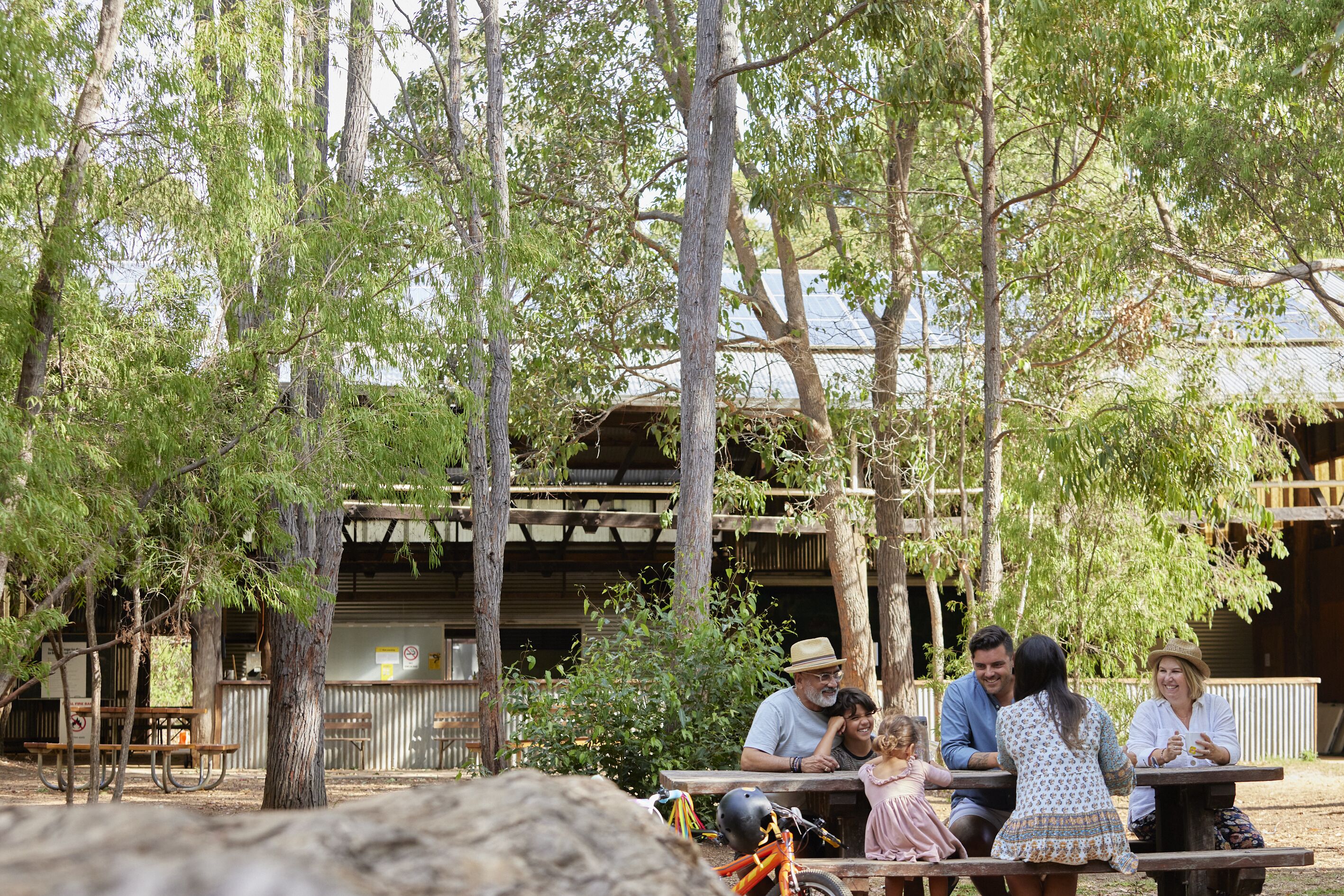 Une famille est assise à une table en bois à l’extérieur, et on aperçoit un bâtiment rustique en arrière-plan.