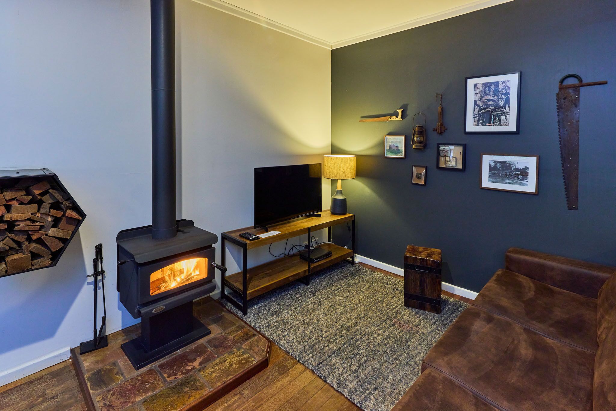 Ein gemütliches Wohnzimmer mit Kaminofen, Fernseher auf einem Holzständer, einer Lampe und gerahmten Bildern an der Wand.