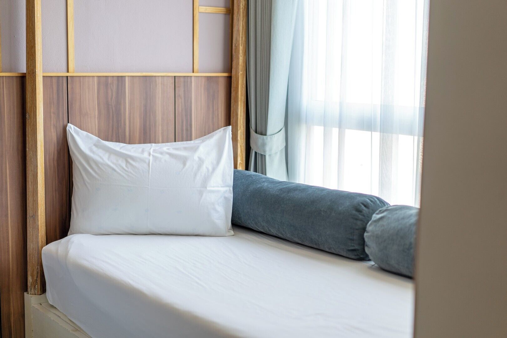 A neatly made bed with white bedding and a grey pillow, set against a wooden headboard and a window with sheer curtains.