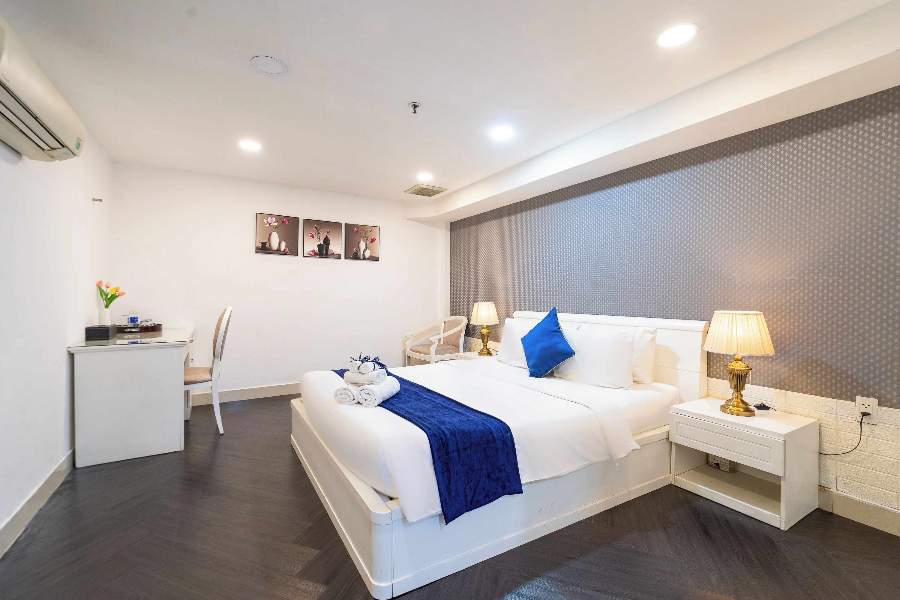 A modern hotel room with a neatly made bed, a bedside table with a lamp, a desk with a chair, and a wall-mounted air conditioner.