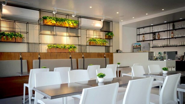 Un comedor moderno con sillas y mesas blancas, un separador de madera, y estanterías con plantas y cristalería.