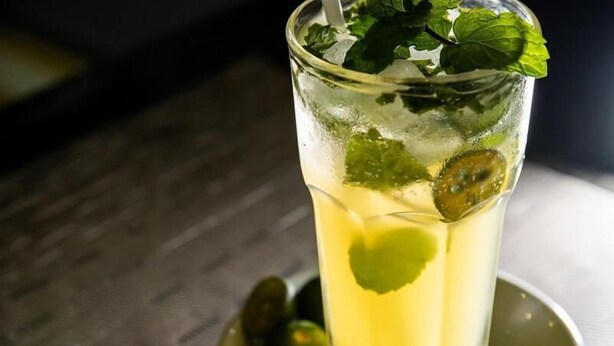 Un vaso de bebida amarilla con hojas de menta y una guarnición con forma de corazón, servido en un plato con una cuchara.
