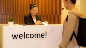 Un mostrador de recepción con un cartel que dice '¡bienvenidos!', una planta en maceta y una persona sentada detrás.