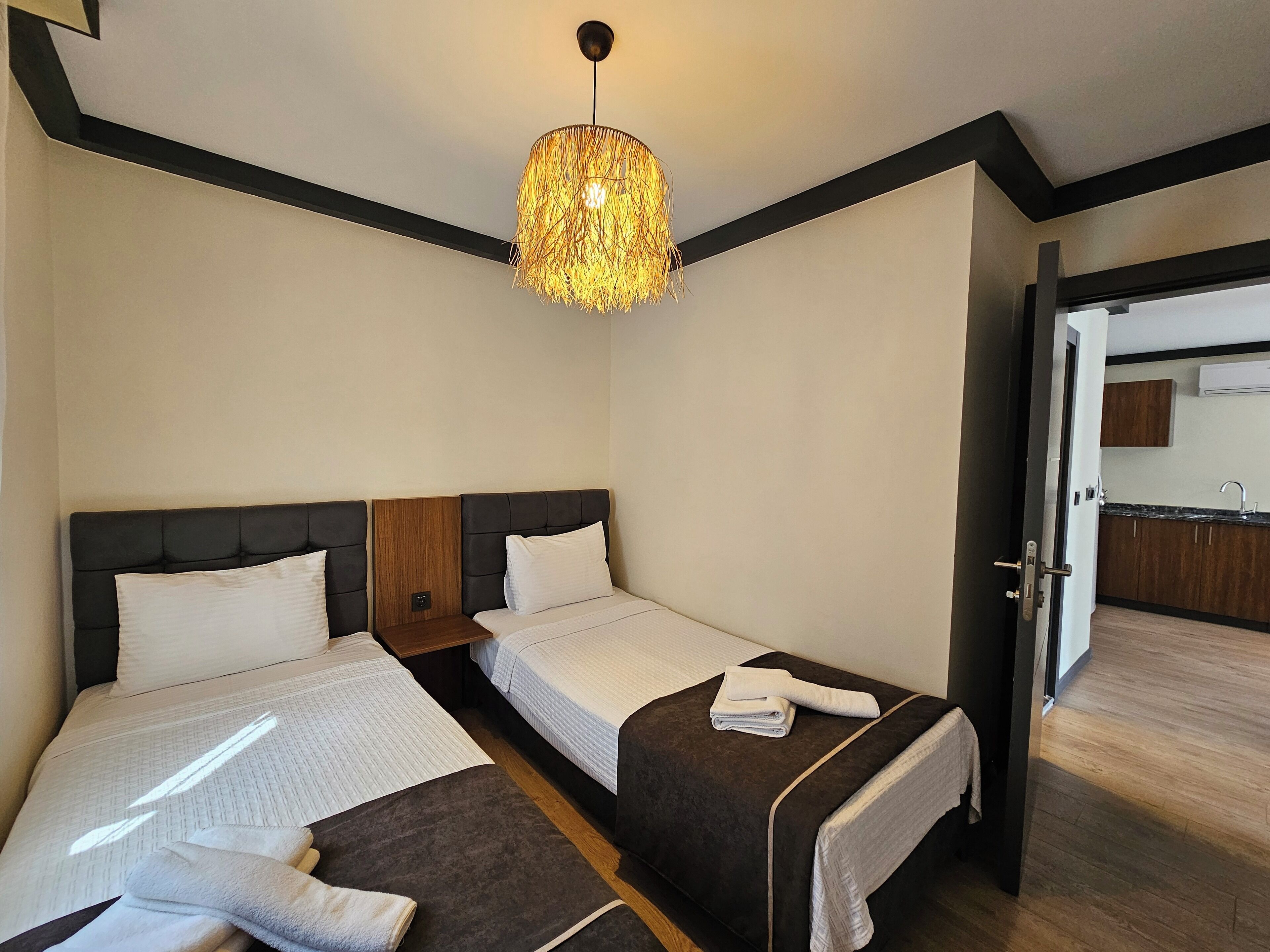 A hotel room with two beds, a chandelier, a wooden headboard, a small table, and a view of the kitchen through an open door.