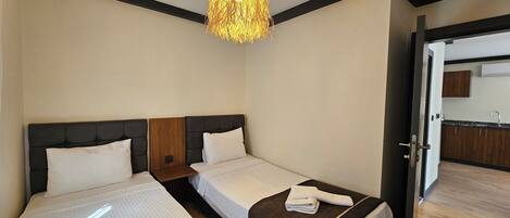 A hotel room with two beds, a chandelier, a wooden headboard, a small table, and a view of the kitchen through an open door.