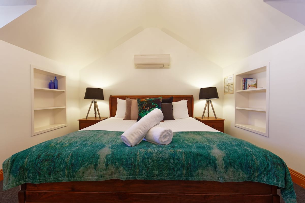 A neatly made bed with a green blanket, two bedside lamps, and a wall-mounted air conditioner.