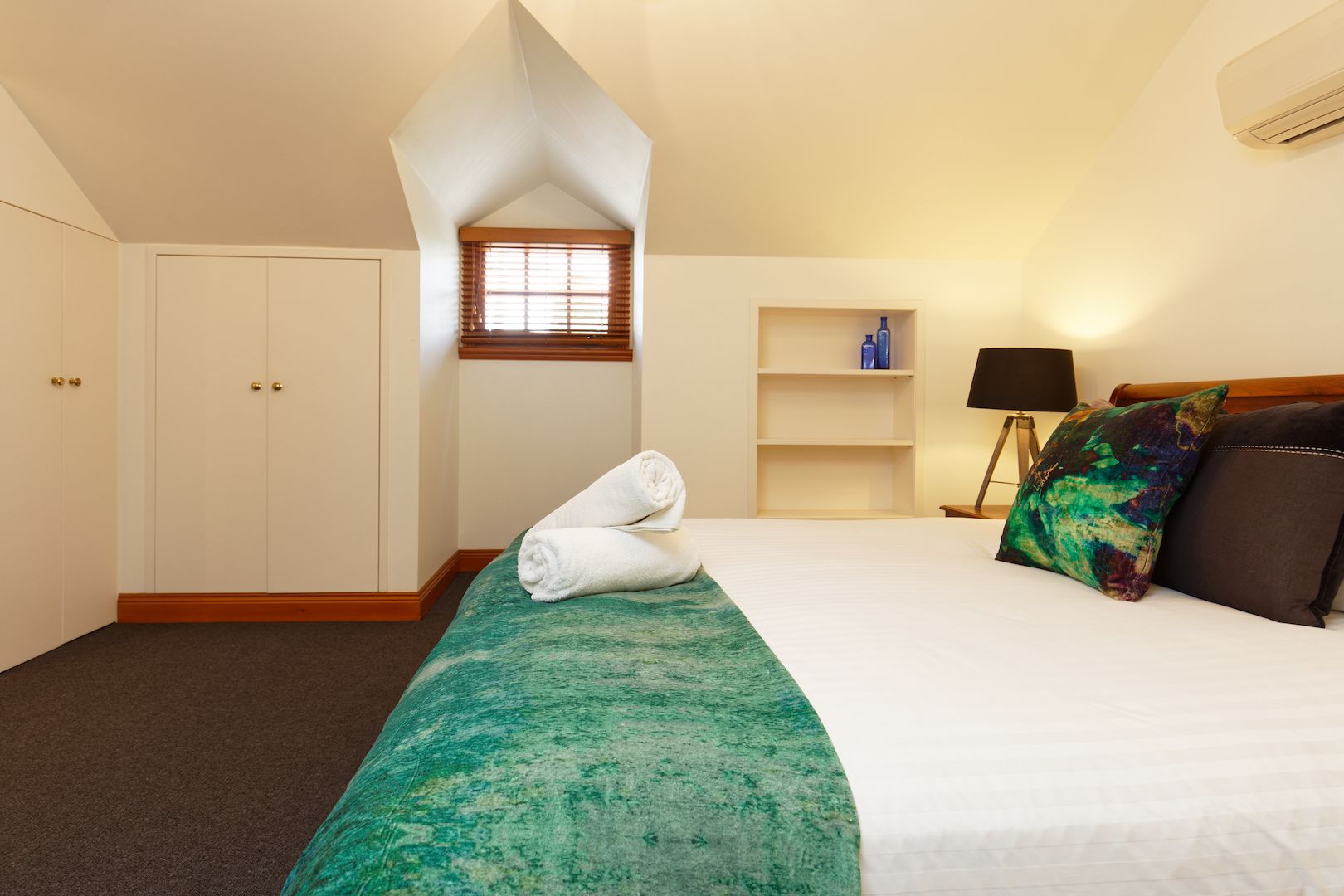 Une chambre d’hôtel avec un lit, une fenêtre avec des stores, une table de chevet avec une lampe et une étagère avec des articles de toilette.