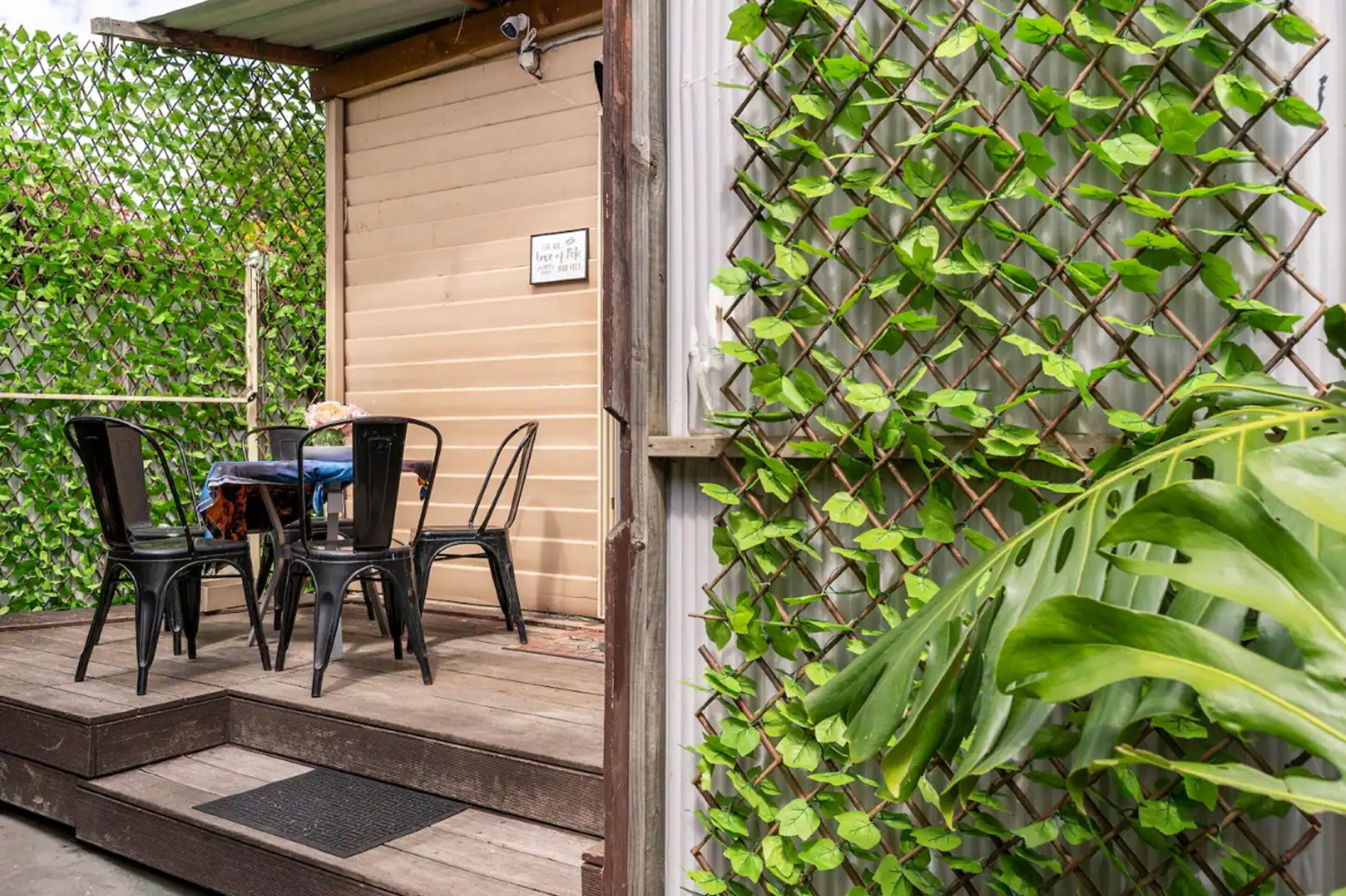 A covered outdoor seating area with metal chairs and a table, a wooden door, and a lattice fence with green foliage.
