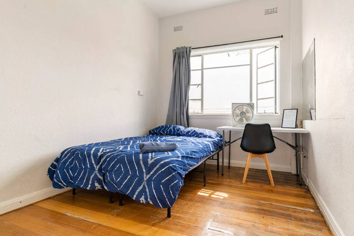 Una habitación pequeña y ordenada con una cama individual, un escritorio con silla, un ventanal con cortinas y un ventilador de pie.