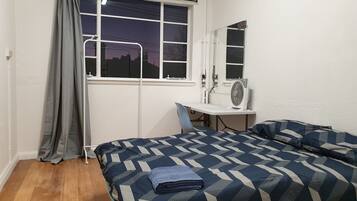 A bedroom with a bed, a desk, a chair, a window with curtains, and a mirror.