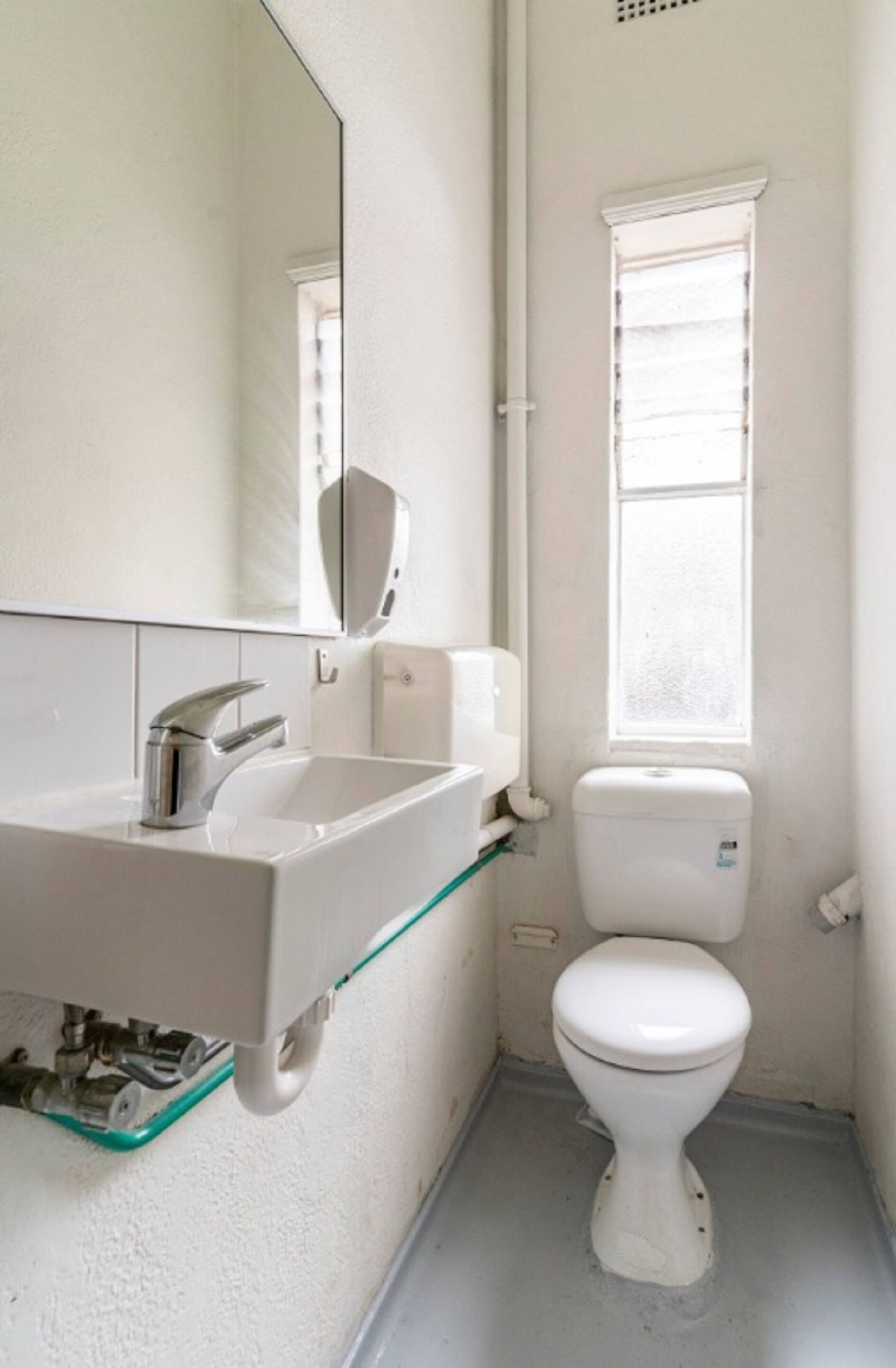 A small, compact bathroom with a white sink, a mirror, a toilet, and a window.