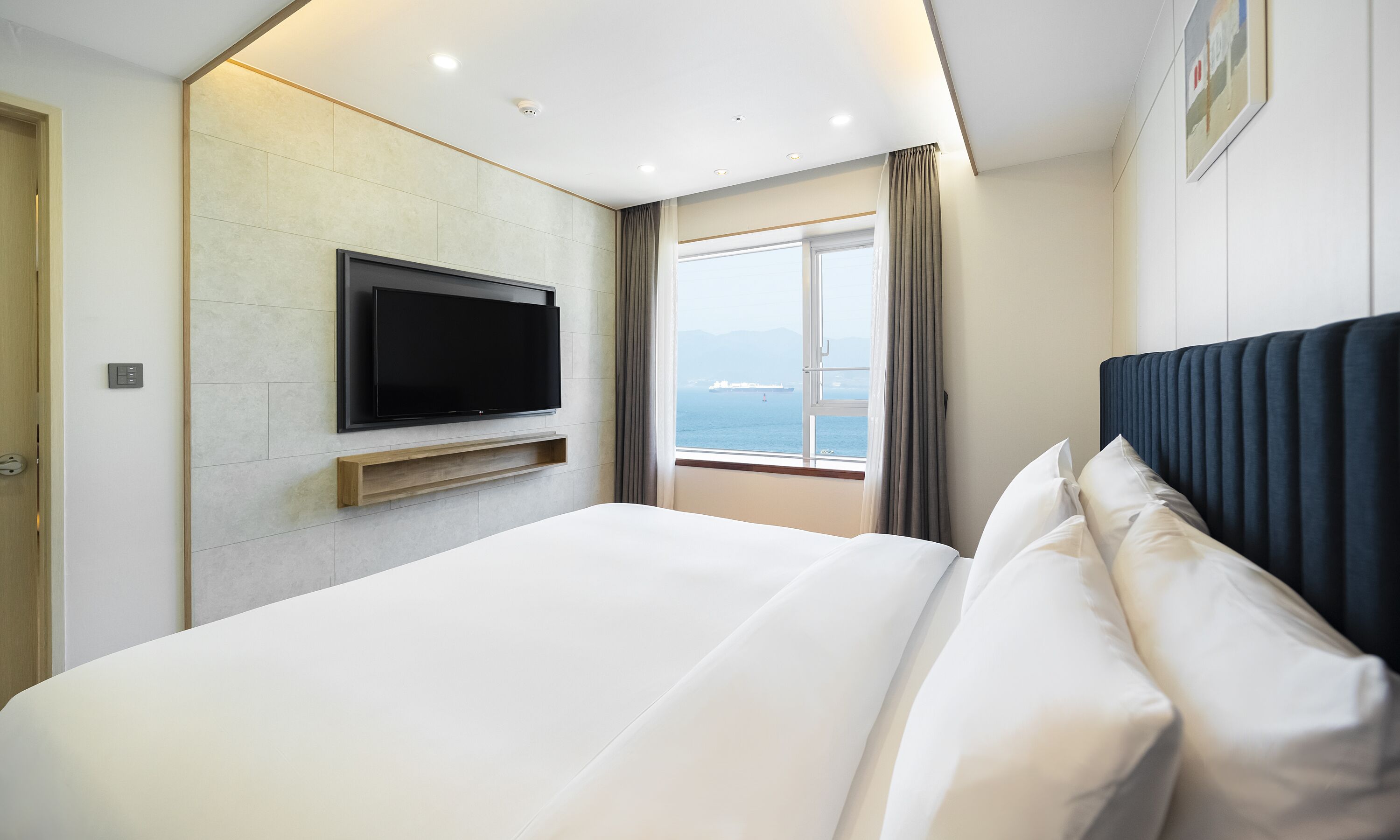 A modern hotel room with a large bed, a flat-screen TV mounted on the wall, and a window offering a view of the sea.