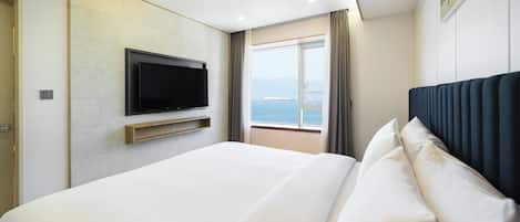 A modern hotel room with a large bed, a flat-screen TV mounted on the wall, and a window offering a view of the sea.