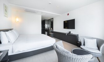 Una habitación de hotel moderna con una cama grande, una pequeña zona de estar con una silla y una mesa, un televisor de pantalla plana montado en la pared y una kitchenette con un lavamanos y un espejo.
