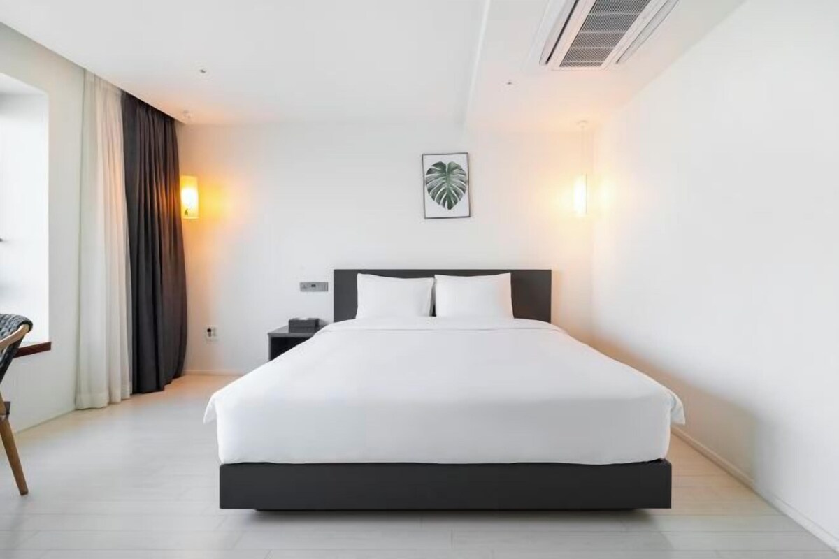 A modern hotel room with a large bed, a nightstand, a chair, and a wall-mounted light.