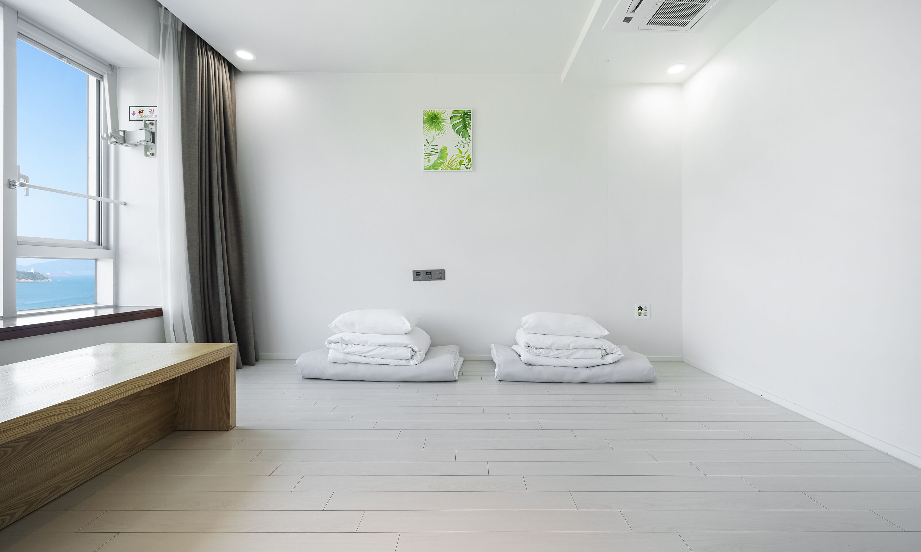 A minimalist hotel room with a wooden bench, two neatly folded white towels, a window with a view of the sea, and a green plant on the wall.