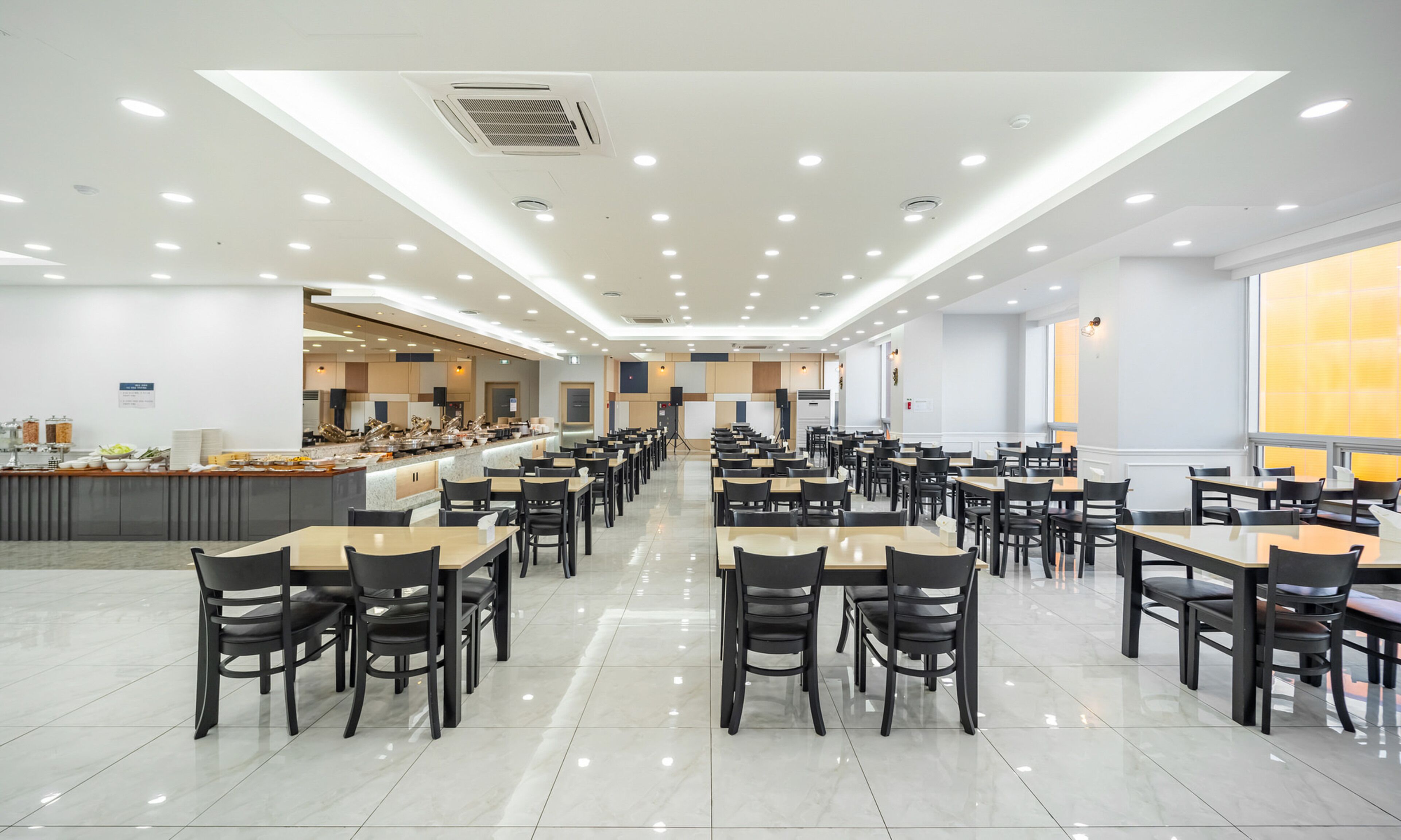 A spacious dining area with multiple tables and chairs, a buffet counter, and a bright, well-lit ceiling with recessed lighting.