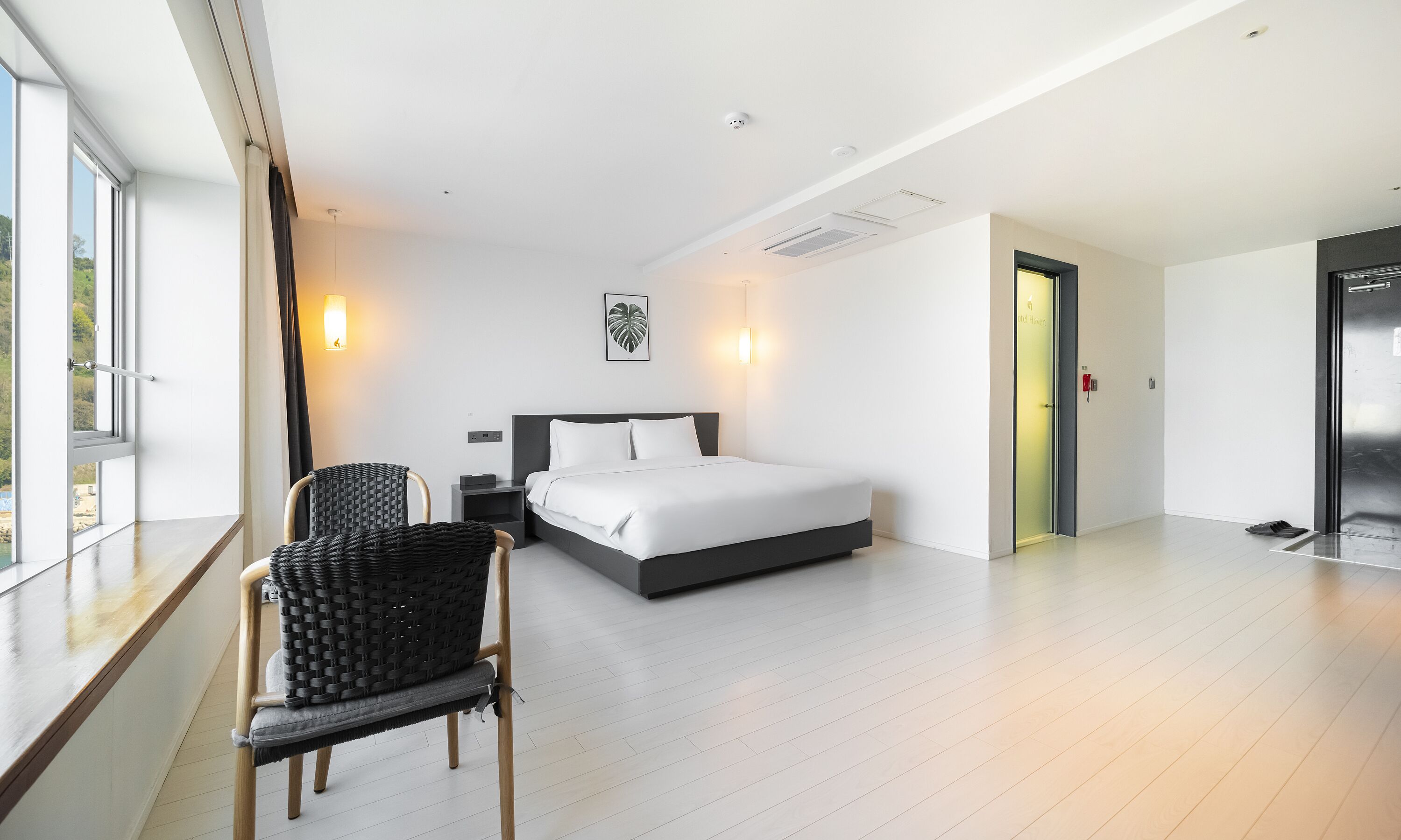 A modern hotel room with a large bed, a seating area with a chair, and a view through the window.