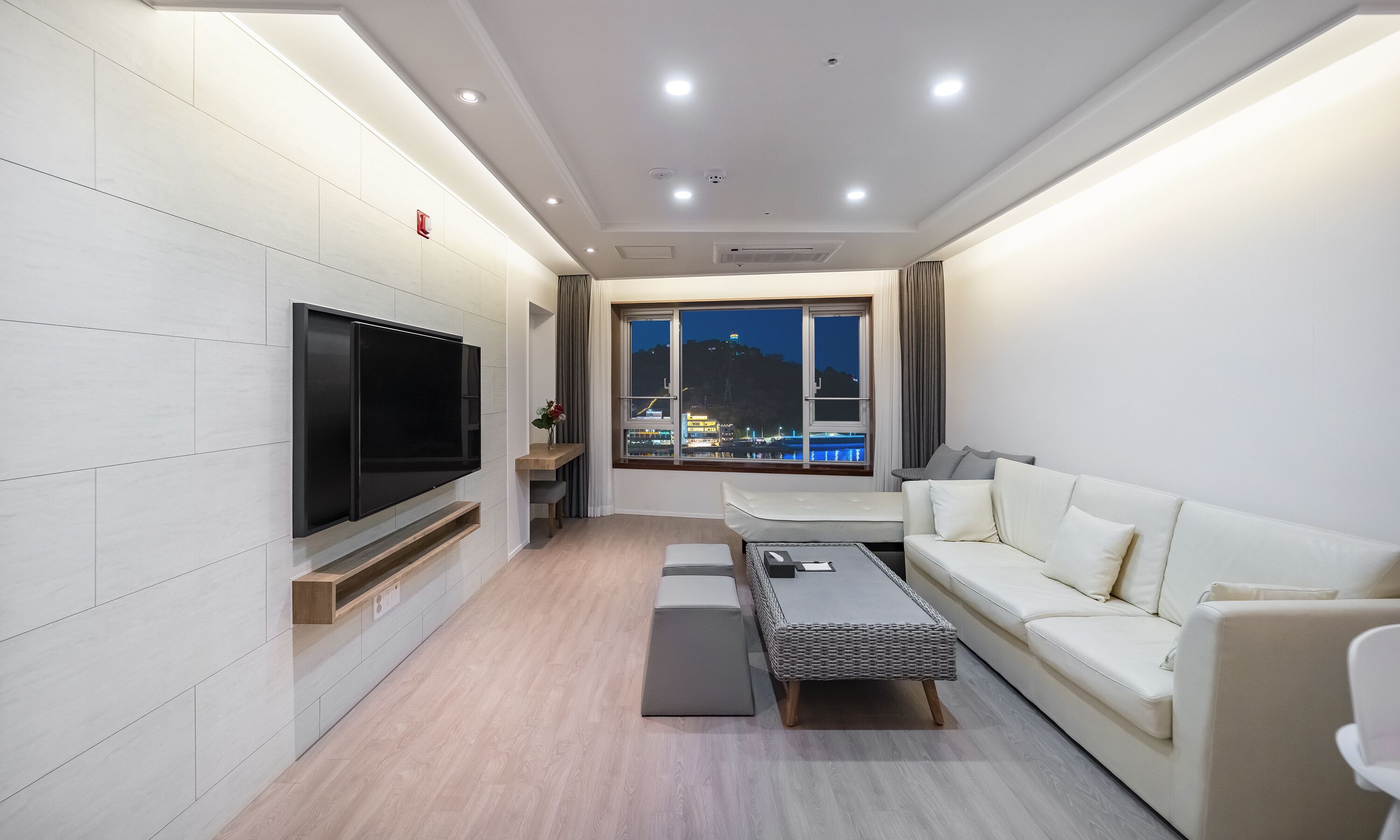 A modern living room with a flat-screen TV, a sofa, a coffee table, and a window with a view of a cityscape at night.