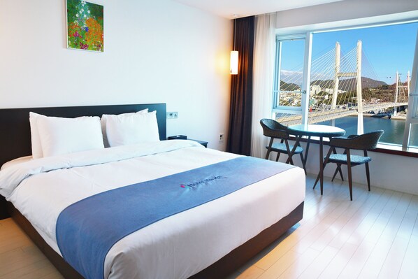 A hotel room with a large bed, a dining area with a table and chairs, and a view of a bridge and water. - Hotel Haven (Yeosu)