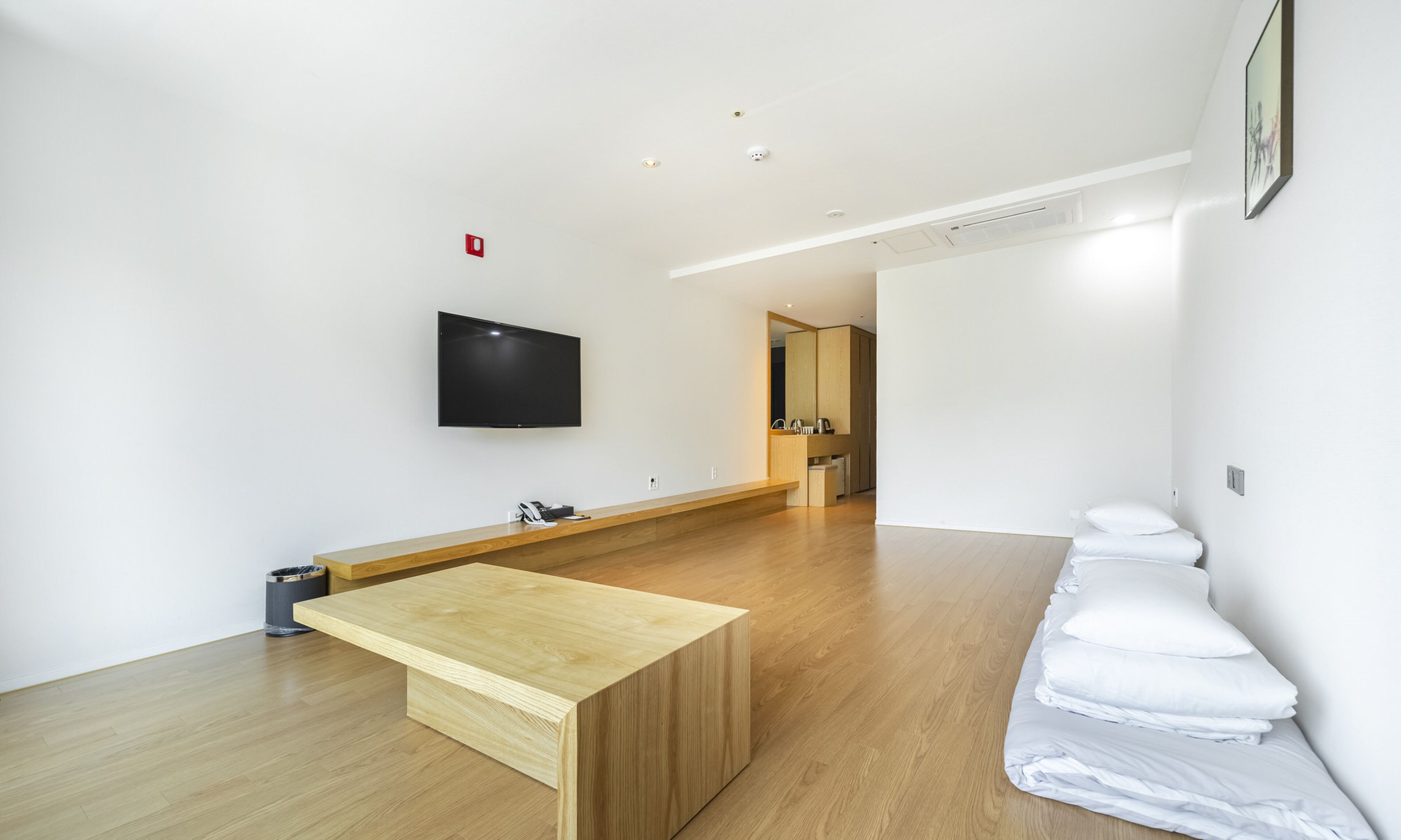 Quarto de hotel moderno com piso de madeira, TV de tela plana na parede, um banco de madeira e uma cama bem arrumada com lençóis brancos.