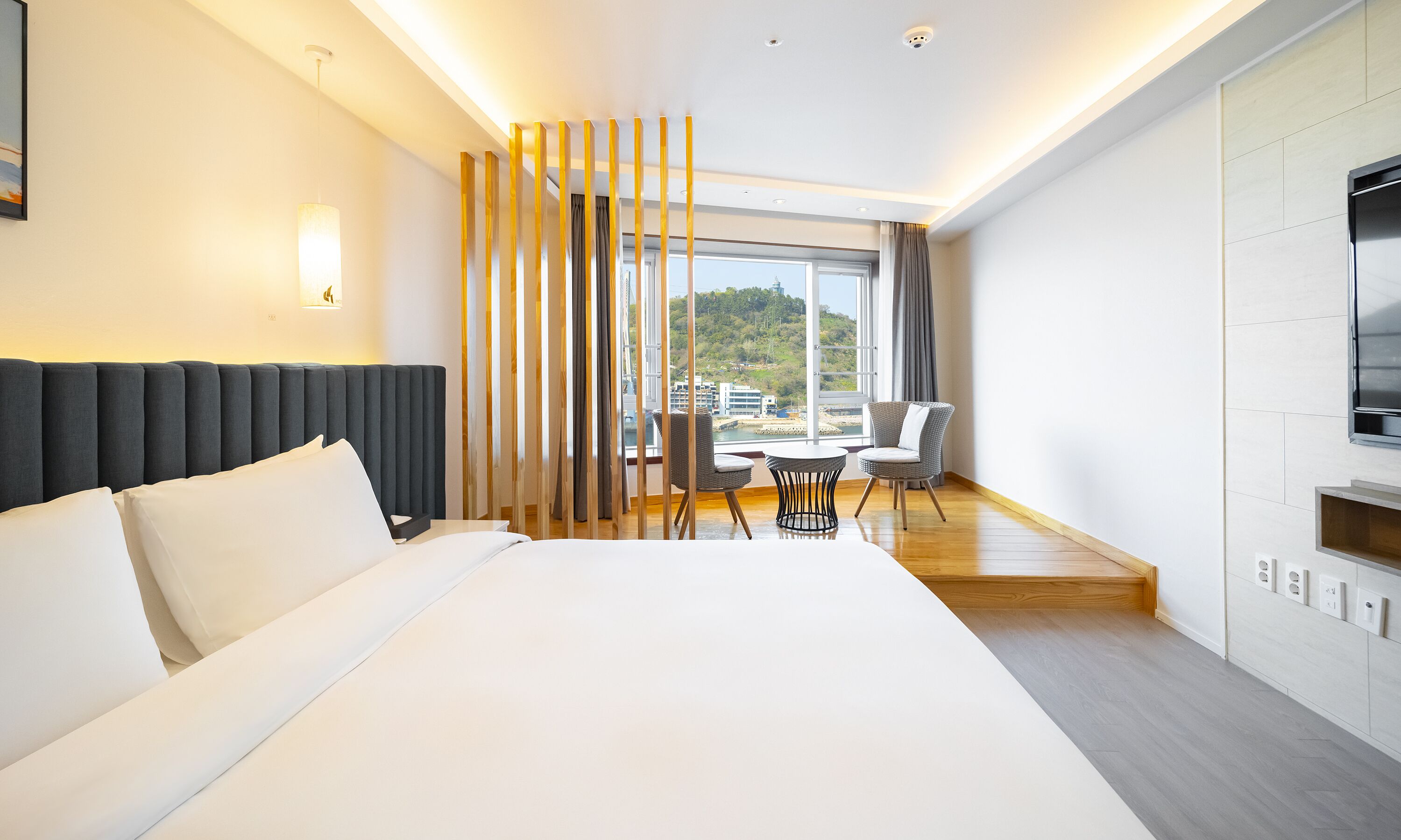 A modern hotel room with a large bed, a dining area with two chairs, a view of the city, and a TV mounted on the wall.