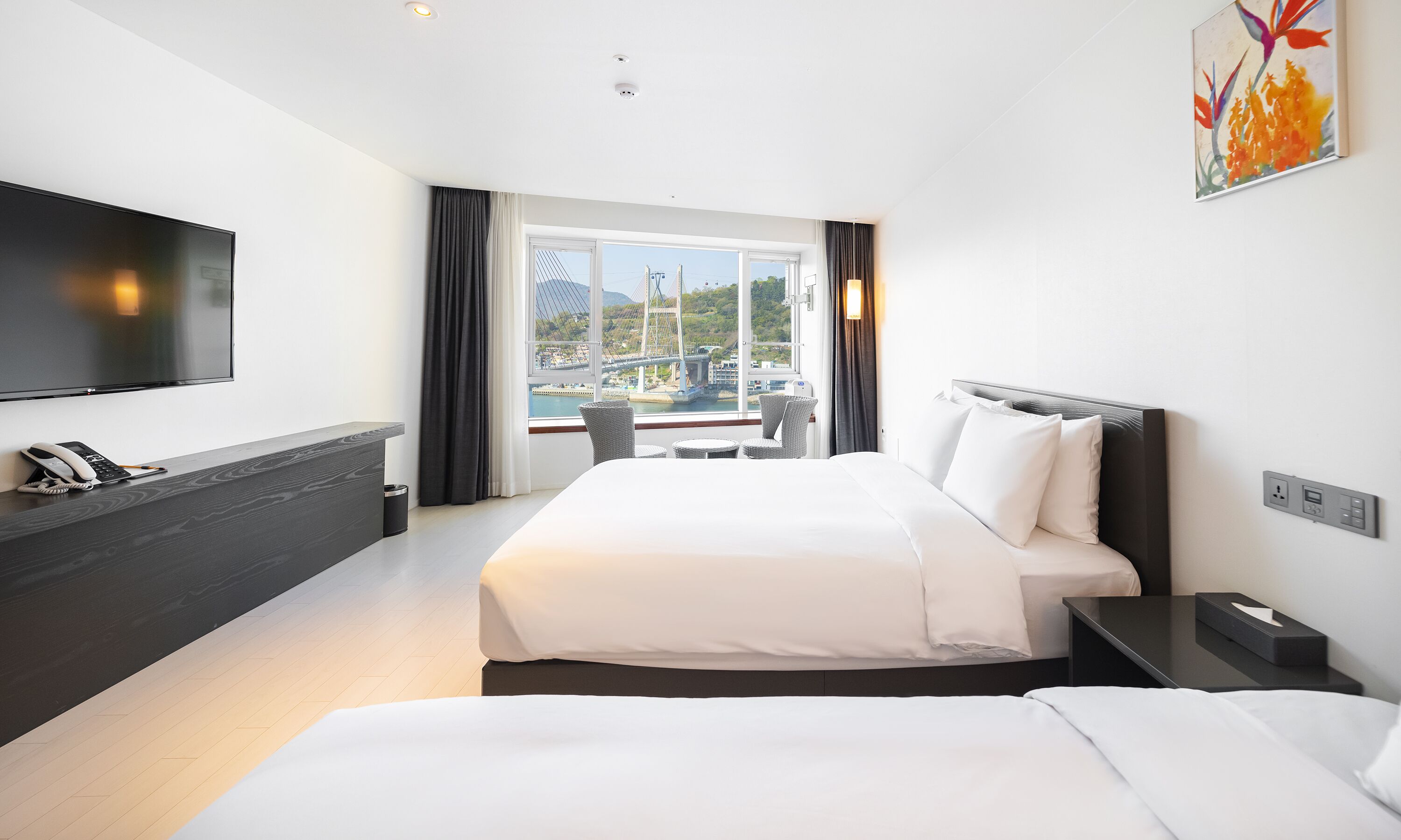 A hotel room with a bed, bedside tables, a TV, a phone, and a window with a view of a bridge and greenery.