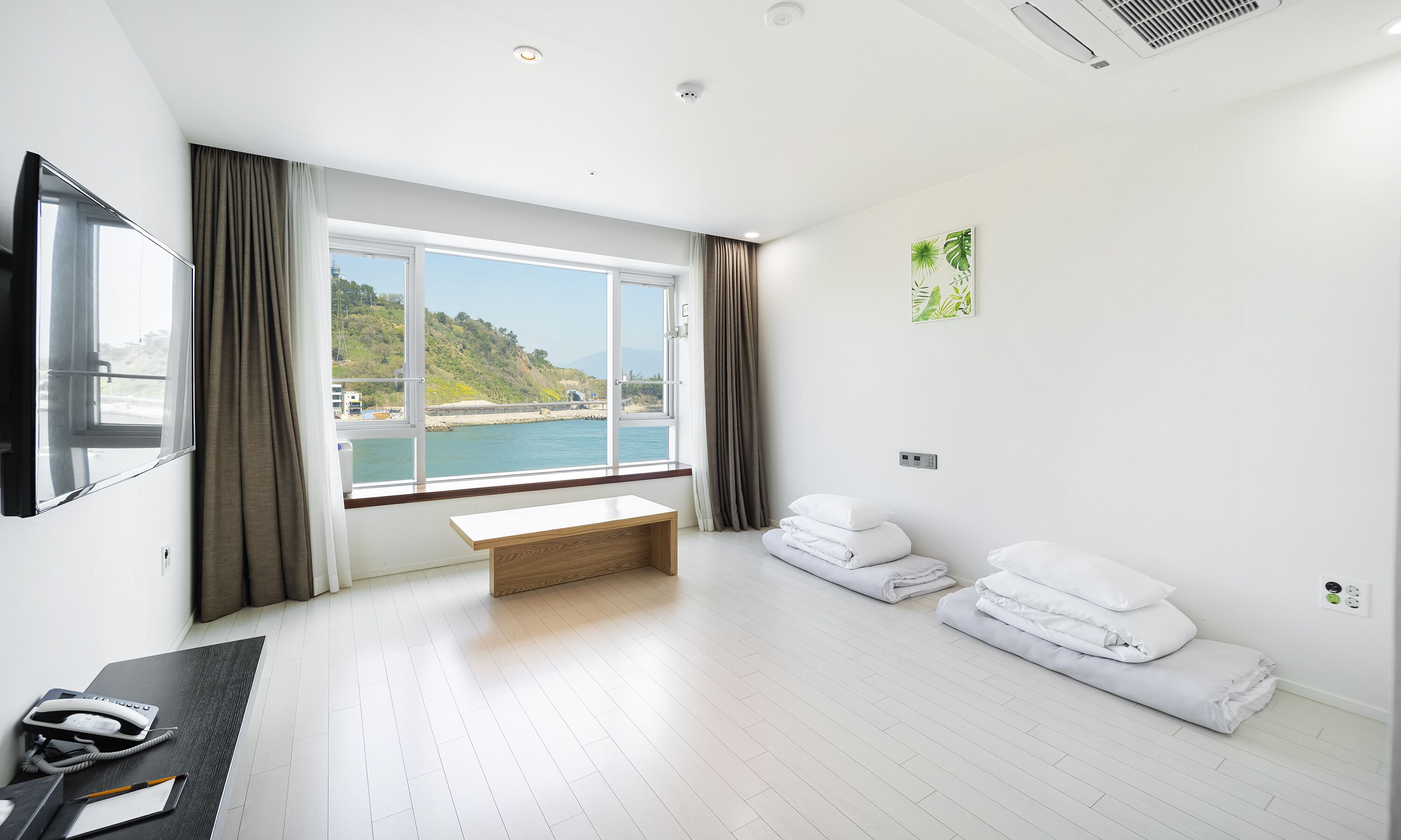 A hotel room with a large window, a bed with white linens, a bedside table, and a desk with a telephone and notepad.