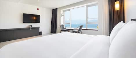 A hotel room with a large bed, a desk with a telephone, a TV mounted on the wall, and a window offering a view of the sea.