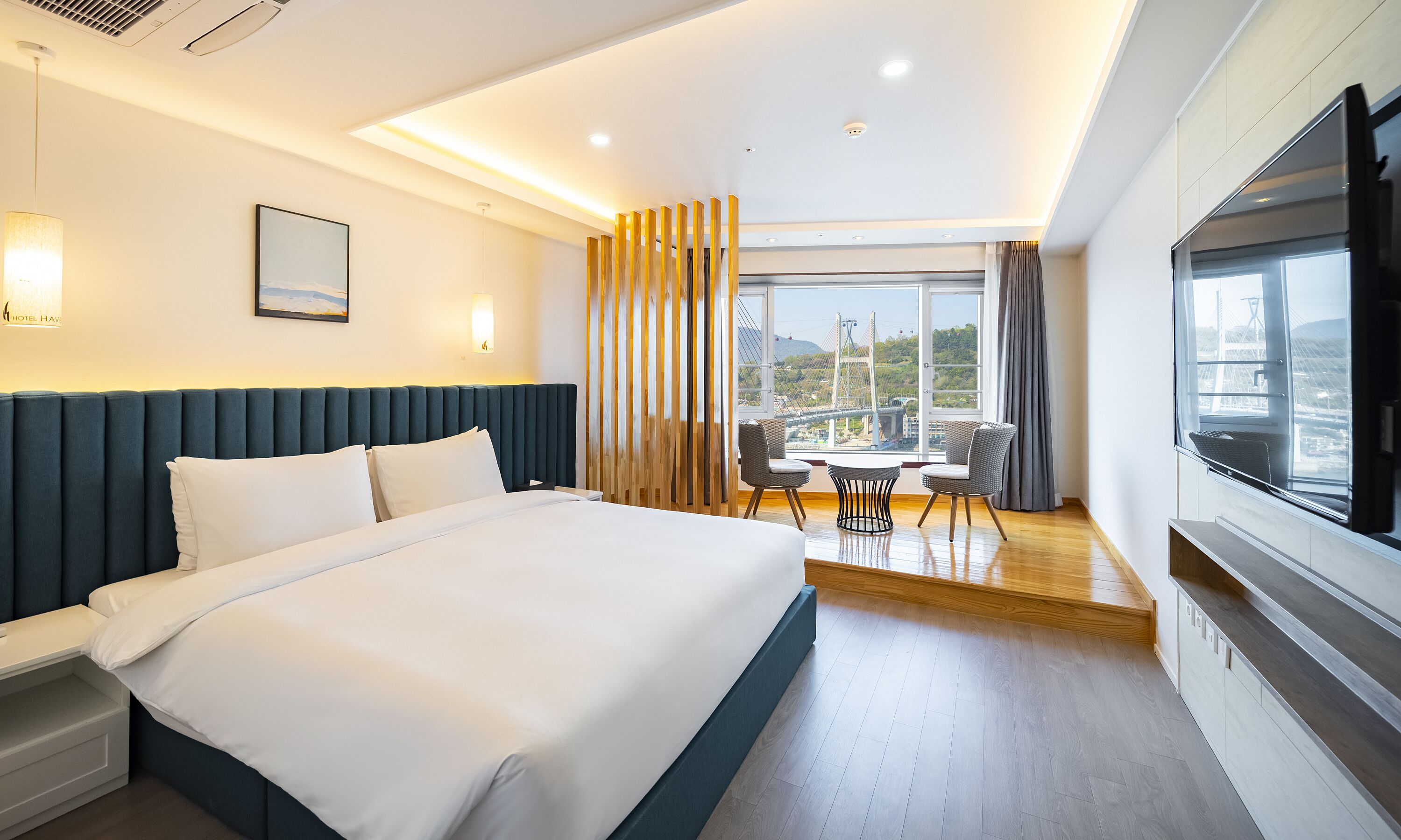 A hotel room with a large bed, a dining table with chairs, a TV, and a view of a bridge.