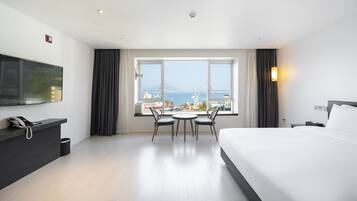 Een hotelkamer met een bed, een bureau met telefoon, een televisie, een raam met uitzicht op zee en gebouwen, en een tafel met stoelen.