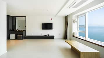 A modern hotel room with a large window, a flat-screen TV mounted on the wall, a wooden bench, and a minimalist bathroom with a large mirror and a sink.