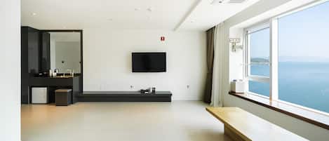 Una habitación de hotel moderna con un ventanal grande, un televisor de pantalla plana montado en la pared, un banco de madera y un baño minimalista con un espejo grande y un lavabo.