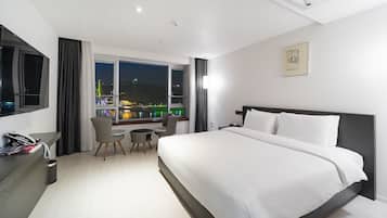Habitación de hotel con una cama grande, un escritorio con silla, un teléfono y una ventana que ofrece vistas de la ciudad de noche.