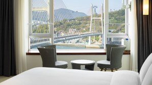 Un cuarto de hotel con una cama, dos sillas de mimbre, una pequeña mesa redonda y un amplio ventanal con vistas a un puente, edificios y follaje.
