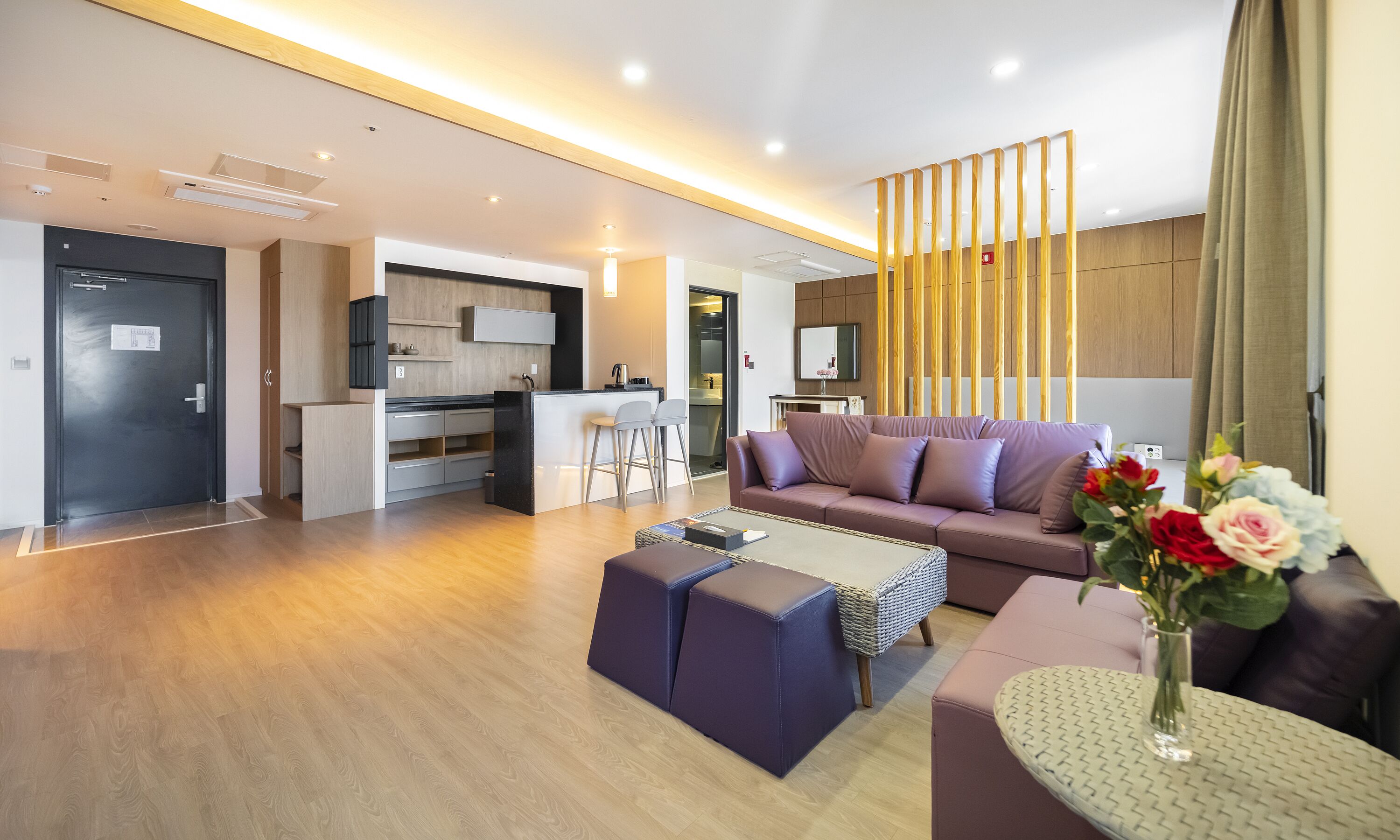 Una sala moderna con sofá, reposapiés y mesa de centro, un área de cocina con taburetes de bar, y una puerta que da a un baño.