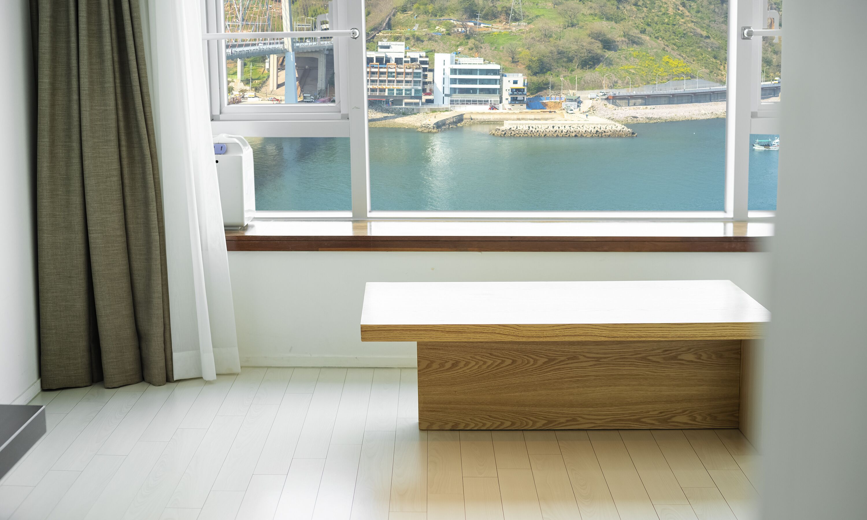 Una habitación con un banco de madera junto a una ventana con vista al puerto, piso blanco y vista a edificios y un puente.