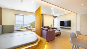 Habitación de hotel moderna con una cama grande, zona de estar con sofá y sillones, televisor montado en la pared y ventana con vistas a la ciudad.