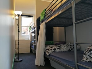 A dormitory room with bunk beds, a standing lamp, a window, and a radiator. - Landing Pads Brunswick - Hostel (Brunswick)