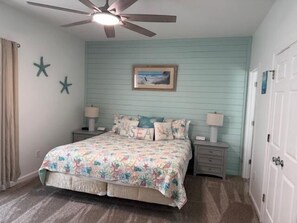 A bedroom with a bed, two bedside tables, a ceiling fan, a wall-mounted picture, and a door.