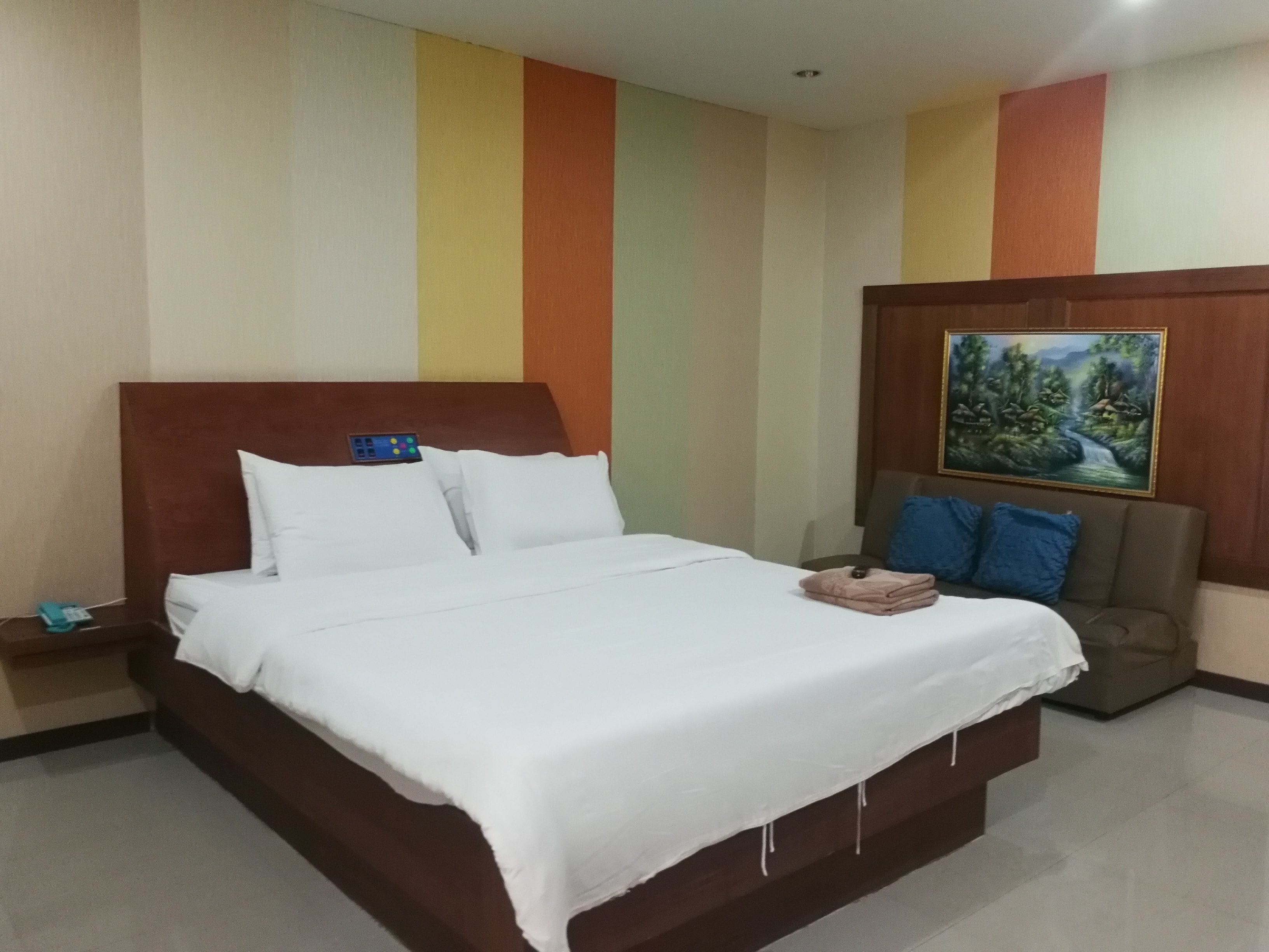A hotel room with a large bed, a nightstand, a chair with blue cushions, and a painting on the wall.