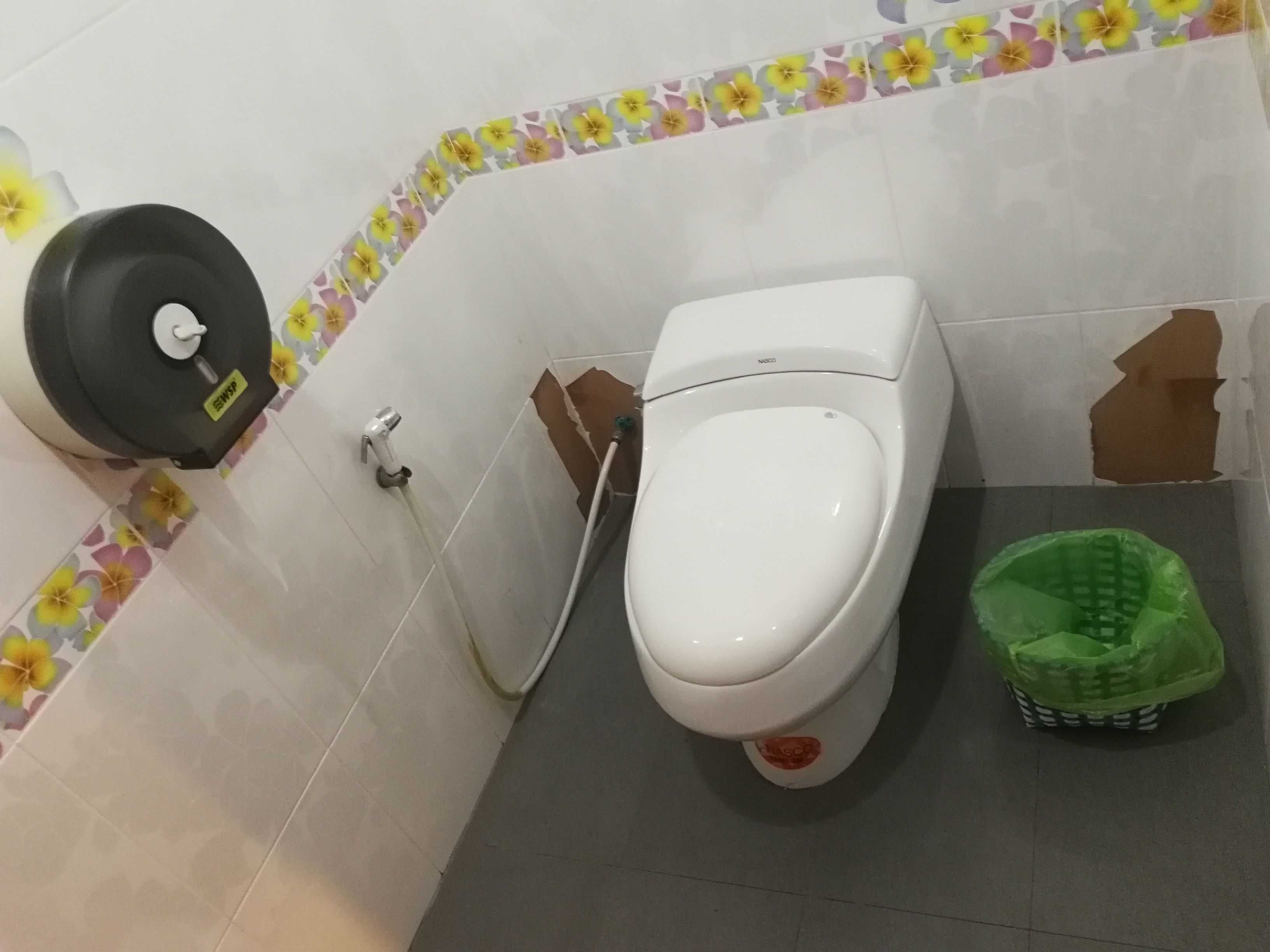 A toilet with a hand sanitizer dispenser, a green trash bin, and a damaged wall.