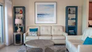 Uma sala de estar com um sofá bege, uma mesa de centro redonda de madeira e um quadro de paisagem na parede.