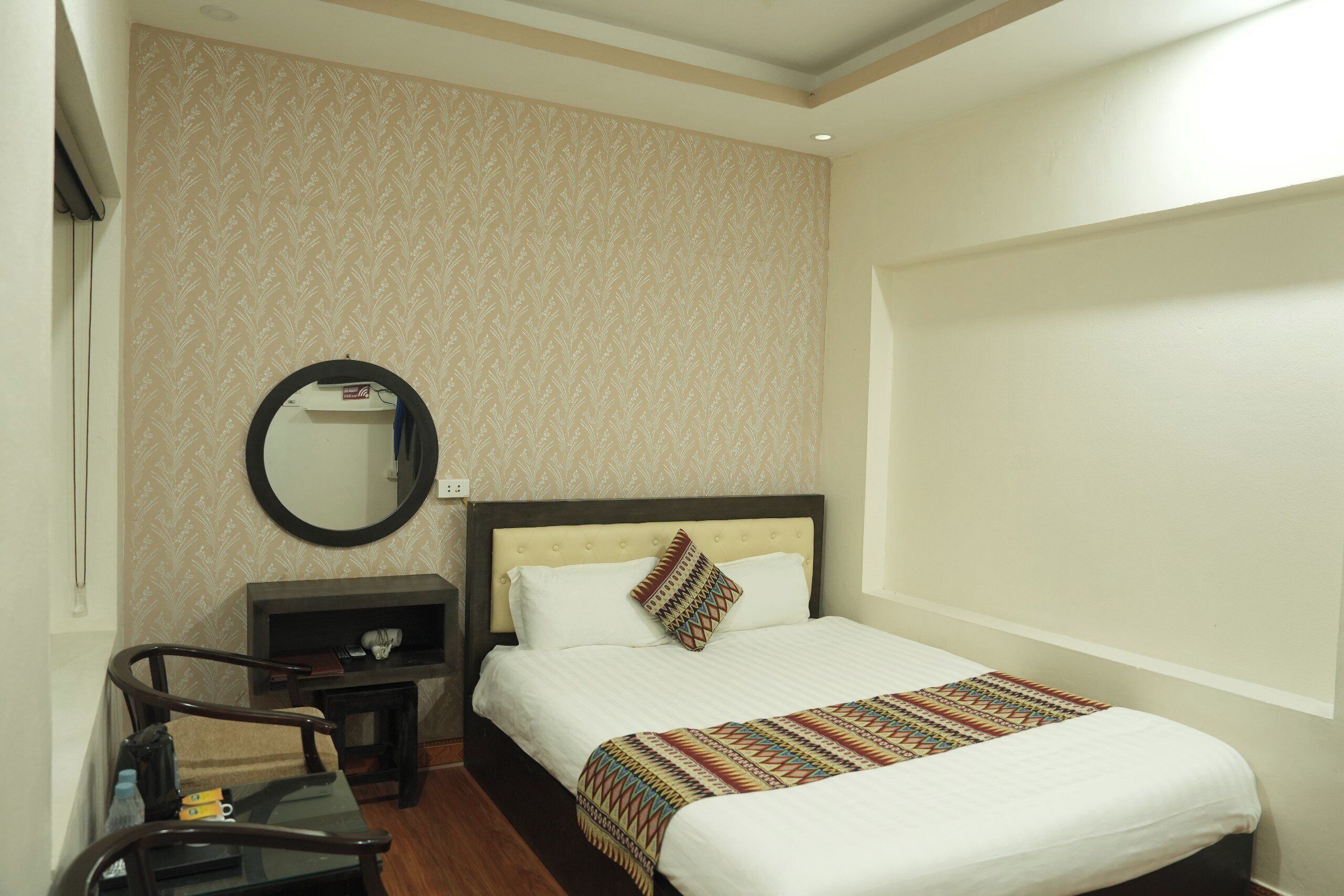 A hotel room with a bed, a bedside table, a mirror, and a small desk with a chair.