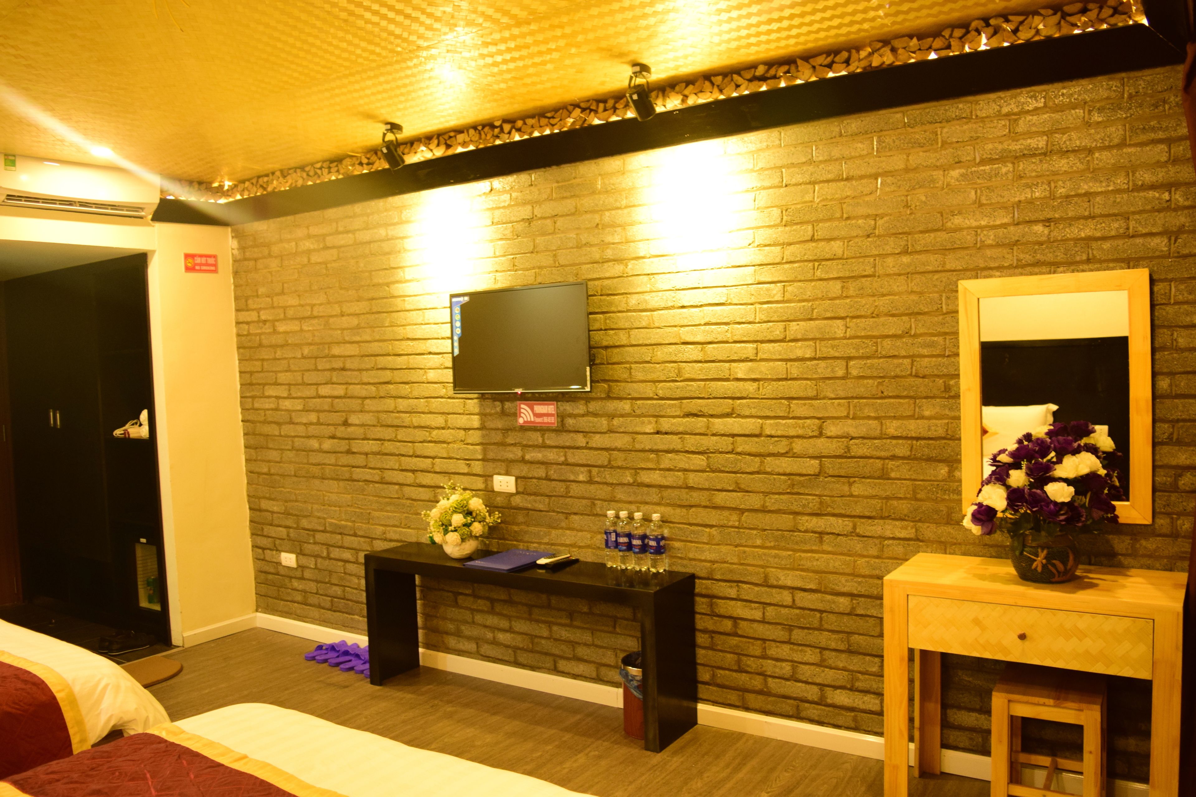 Ein Hotelzimmer mit Backsteinwand, einem flachbildfernseher über einer Holztruhe und einem Bett mit roter Bettwäsche, sowie einem kleinen Holztischchen mit Blumenvase.